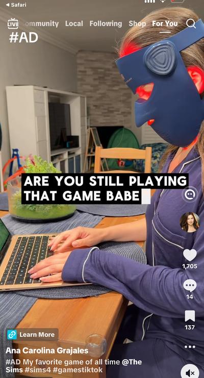 Ana wearing LED face mask at laptop with text 'Are you still playing that game babe' — The Sims 4 ad